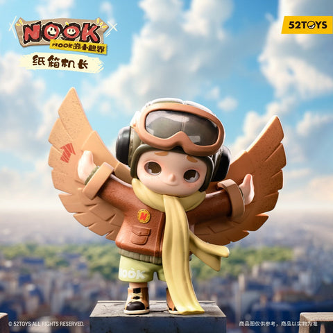 A small pilot-themed figurine stands on a rooftop ledge with cardboard wings spread wide behind their arms. The child wears a brown jacket, yellow scarf, green shorts, and a large aviator helmet with goggles. A bright blue sky with soft clouds and a blurred cityscape fills the background below.