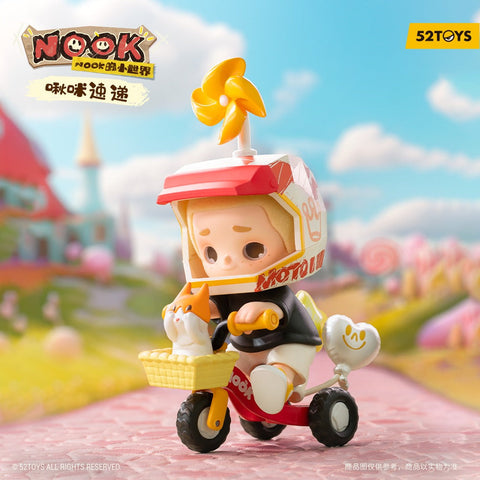A child figurine rides a small three-wheeled scooter along a pastel path, holding the handlebars while an orange-and-white cat sits in a front basket. The child wears a boxy helmet topped with a spinning yellow pinwheel. A candy-colored village with whimsical houses and trees blurs in the bright background.