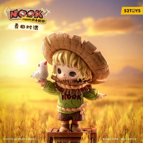A scarecrow-themed child figurine stands on a wooden crate in a golden wheat field at sunset. The character wears a large woven hat, green sweater with Nook lettering, brown shorts, and a straw-like ruff around the neck. A small white bird perches on one outstretched hand as warm light fills the sky.