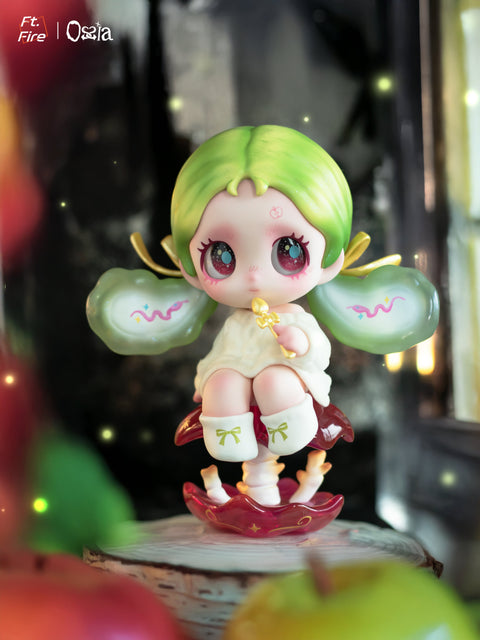 A vinyl figure with lime-green hair and clear wing-like extensions holds a small yellow object while sitting on a red crescent-shaped base. Fireflies and blurred fruit fill the softly lit background, creating a dreamy natural setting.