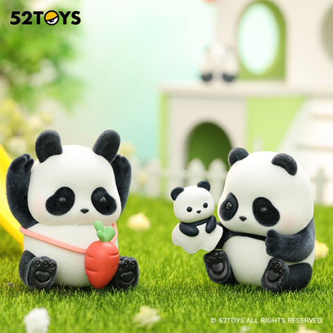 Two panda figurines sit in a grassy playground setting. One raises its arms while holding a carrot-shaped toy with a pink strap. The other holds a tiny panda figure in its lap. Both have rounded bodies, black paws, and soft expressions, with pastel play structures behind them.