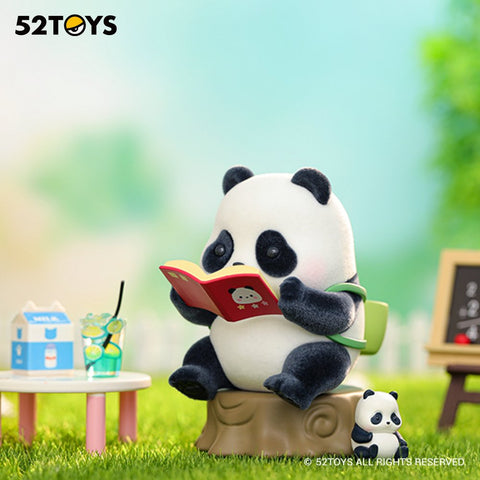 A large panda figurine sits on a small tree-stump seat reading a red picture book in a grassy outdoor play area. A tiny panda figure sits beside it. A white table holds a milk carton and a green drink with lemon slices, while a small chalkboard stands blurred in the background.
