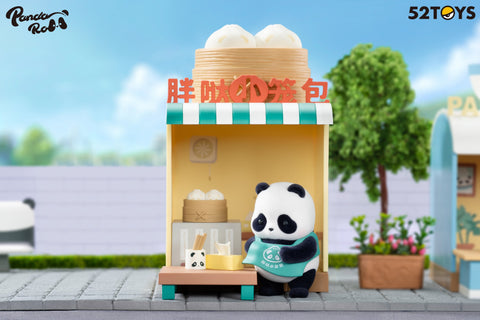 A panda figurine from the Panda Roll Shopping Street series by 52TOYS is shown sitting in front of a small yellow bun shop with a green-and-white striped awning. The panda wears a teal apron and is holding a bamboo steamer basket, preparing steamed buns. The shop features miniature props like chopsticks in a panda-face holder, a basket of buns on a table, and a stove inside. The background includes a tree, flower planter, and part of another storefront, all set against a pastel outdoor scene.