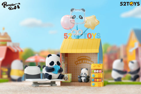 A panda figurine from the 52TOYS Panda Roll Shopping Street series sits on a white-and-black bench reading a blue booklet outside a miniature toy store with a bright yellow roof. The shop features a small gacha-style capsule machine, a seated panda toy figure, and colorful signage with balloons shaped like a bear, star, and circle. In the background, other panda figures and storefronts add to the vibrant and playful shopping street scene set under a sunny sky with fluffy clouds.