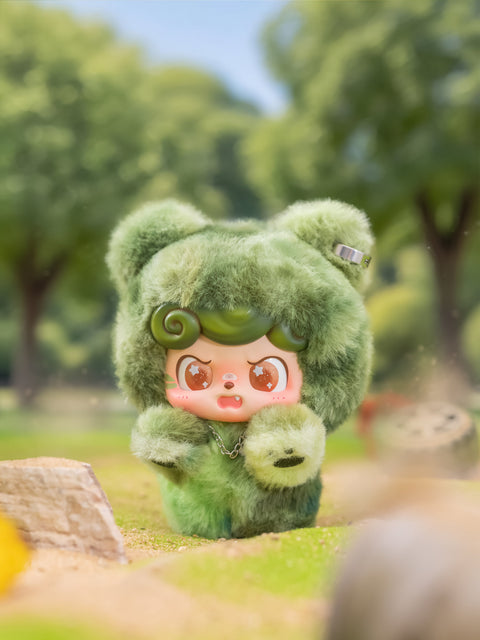 A fuzzy green Qkid plush stands in a grassy park setting with an angry expression. Its bear-ear hood and green fur blend with the trees behind it. The character clenches its tiny paws in frustration, with sunlight filtering softly through the leaves.