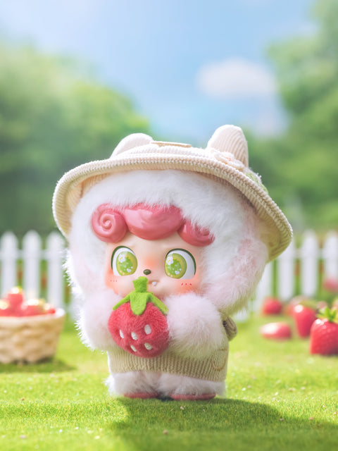 A white plush Qkid character with pink curls holds a large felt strawberry in both hands. It wears a beige hat and overalls, standing in a sunlit garden surrounded by green grass, strawberries, and a white picket fence beneath a bright blue sky.