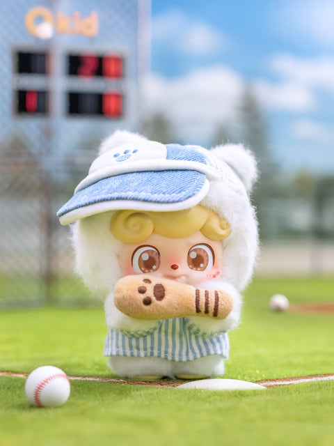 A soft yellow Qkid plush dressed in a fluffy white hood and blue striped overalls holds a plush baseball bat. The figure’s large brown eyes and blush cheeks face forward. A scoreboard and baseball field can be seen behind it under clear skies.