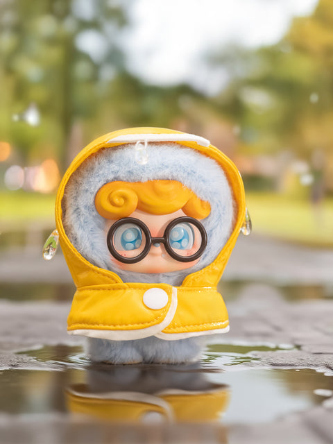 A blue Qkid plush with orange hair and large round glasses stands in a puddle wearing a yellow raincoat. Water droplets rest on the hood as reflections shimmer below. The blurred background shows a park with trees and glowing bokeh light.