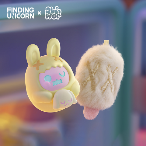 A pair of matching collectibles in soft warm light. On the left is a fuzzy pale beige popsicle plush with stitched X marks and a pink stick. On the right is a small yellow bunny vinyl figure with long ears, wearing a hood marked with stitched X’s and holding a tiny sleeping rabbit plush.