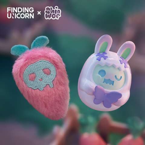 A pair of matching figures float against a blurred green background. The left is a fuzzy strawberry-shaped plush with pink fur and a light blue embroidered skull. The right is a white vinyl bunny figure with purple details and blue-green accents, wearing a hood with small X’s near the ears.
