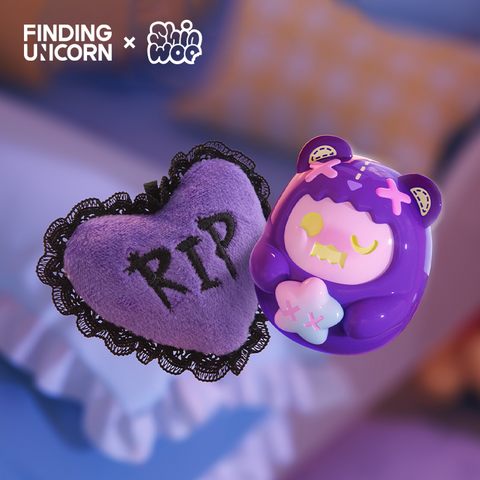 A purple bear-themed vinyl figure floats beside a soft lavender heart-shaped plush with black lace edges embroidered with “RIP.” The bear has yellow and pink facial details, pink X marks on its hood, and holds a white star-shaped pillow. The scene is softly lit with a cozy bedroom backdrop.