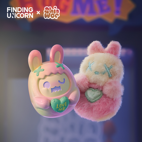 A pastel vinyl bunny figure with pink and mint tones floats next to a fuzzy pink-and-cream plush bunny holding a green heart. The vinyl figure wears a hood with stitched X marks and holds a heart inscribed with “LOVE ME.” A soft sign reading “HUG ME” glows in the background.
