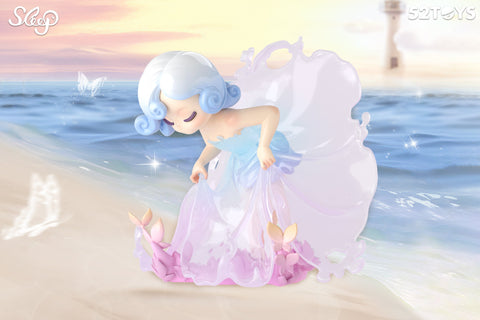 A whimsical 52TOYS Sleep series figurine stands gracefully on a sandy beach at sunset, gazing gently at two small pink sea creatures. The character has curled blue-and-white hair and wears a translucent lavender dress that flows like sea foam. Behind her, calm ocean waves shimmer under the pastel sky, while a lighthouse and glowing butterflies enhance the dreamy seaside ambiance.