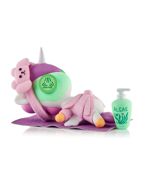 A white Unicorno figure wearing a pink robe and bear sleep mask, lying on a purple mat with a green cucumber slice over its eye next to a bottle of algae lotion.