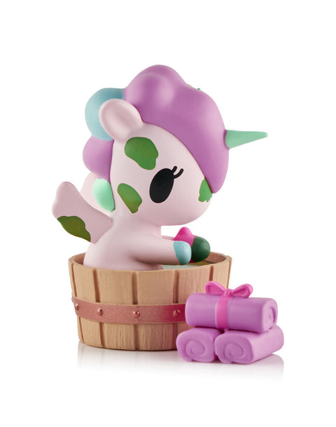 A light pink Unicorno figure with green mud spots sitting inside a brown wooden wash bucket next to a stack of purple rolled towels.