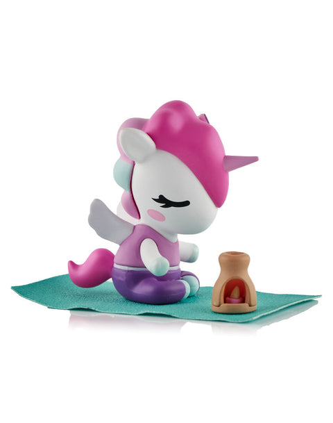 A white and pink Unicorno figure in a seated meditation pose on a teal mat next to a brown incense burner with a pink flame.
