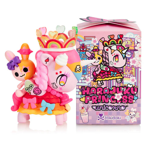 A pink Unicorno figure stands beside its decorative blind box, featuring bright rainbow colors, heart accents, and a translucent crown with candy-like shapes. The figure has pink braids, a multicolored mask, and a small orange rabbit riding on its back. The box shows matching artwork in Harajuku style.