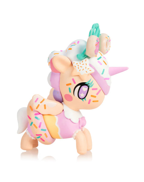 A pastel peach Unicorno decorated with frosting-like white hair is covered in colorful sprinkle shapes. Two small frosted donuts with mint accents rest on its head. It wears a layered skirt with soft yellow and pink tones, matching its wings and hooves. Star markings and gentle colors create a dessert-inspired confection look.