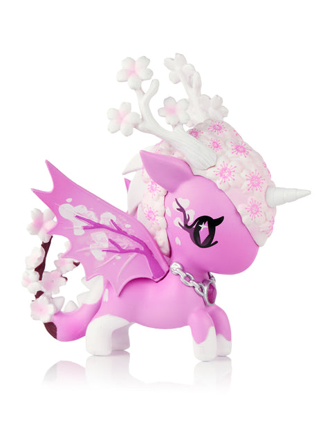 This image shows the Tokidoki Unicorno Mystical Dragon “Cherry Blossom Dragon,” a pink dragon-unicorn figure decorated with white blossom flowers across its head, antlers, wings, and tail. It has a soft matte body, a small horn, and a silver chain collar, posed on a white background.
