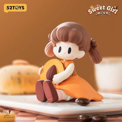 A sweet doll-like figure with auburn pigtails in an orange dress, holding a round almond cookie with a nut center. Her design captures a cozy bakery aesthetic.