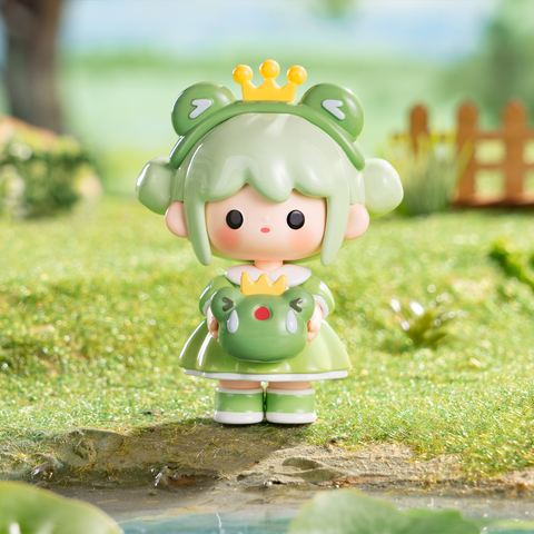 A mint-green haired girl with buns shaped like frog ears wears a green dress and golden crown. She holds a round frog companion also wearing a crown. Standing on grassy ground, this toy captures fairy-tale royalty with whimsical charm and bright pastel fantasy vibes in collectible detail.