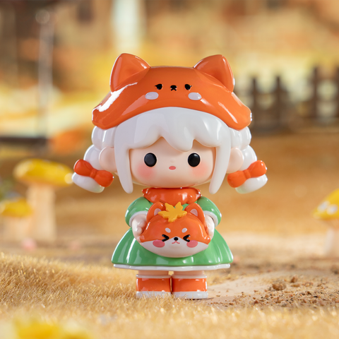 A white-haired figure wears a bright orange fox hood with matching shoes and a green dress. She holds a small round fox companion with perked ears and orange accents. The autumn woodland backdrop enhances the playful forest charm and fabled fox spirit inspiration of this collectible.
