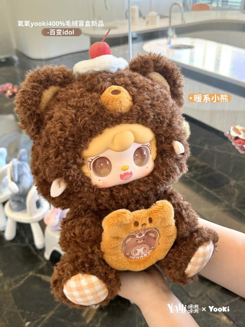 A plush bear doll with curly brown fur and a warm tone sits in hand. The bear has a cherry-topped whipped cream hat, yellow-orange gingham paw pads, and a plush stomach pocket with an embroidered bear face inside, giving a cozy dessert-inspired charm.