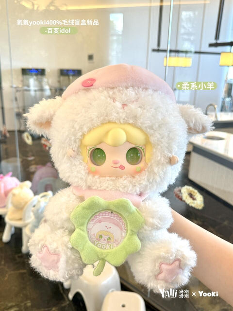 A plush lamb character with fluffy white fur and a pale pink hat is held up indoors. The doll has green eyes, soft blonde hair, and a flower badge with the Yooki logo on its belly. Its hooves feature pink hearts, and its pastel outfit feels cozy and gentle in tone.