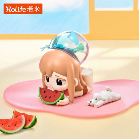 A girl lies flat on her belly with a slice of watermelon in her hands, resting on a pink mat. She wears a blue dome-shaped water hat and has a tiny white puppy companion lounging beside her. The sculpt captures the cooling joy of summer snacking, relaxation, and childhood daydreams in playful form.