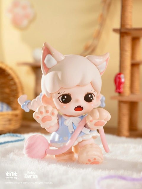 A pale kitten-like figurine with cream ears and a blue outfit kneels on white carpet, holding a pink yarn ball with both paws raised in surprise. The figure has big round eyes, small fangs, and a soft pastel background with wicker furniture and a cat tree behind it.