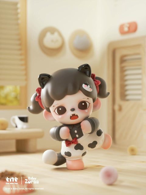 A black-and-white cat-costumed figure with twin pigtails strikes a playful pose on wooden flooring. It wears cow-print pants, a red bow, and glossy black cat ears. Behind it are framed cat silhouettes and soft beige decor matching the cozy tone.