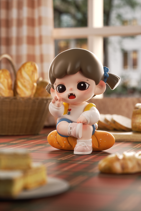 A chibi-style girl figurine with brown pigtails and a blue bow sits cross-legged on an orange baguette-shaped cushion. She wears a white shirt with a heart and “Z” print, blue shorts, and white shoes. The background shows a cozy kitchen scene with bread baskets and soft window light.