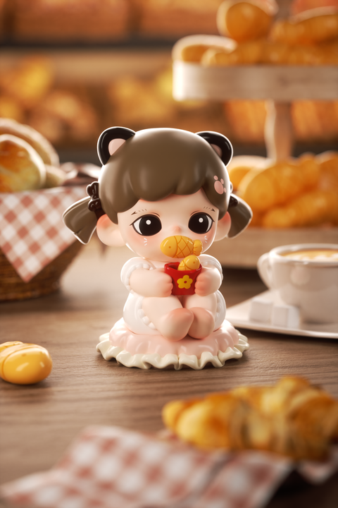 A figurine of a girl with brown hair in pigtails and black cat ears sits on a white ruffled base. She wears a soft white outfit and holds a red cup with small golden fish-shaped pastries. Behind her are croissants, a coffee cup, and a checkered napkin in warm bakery lighting.