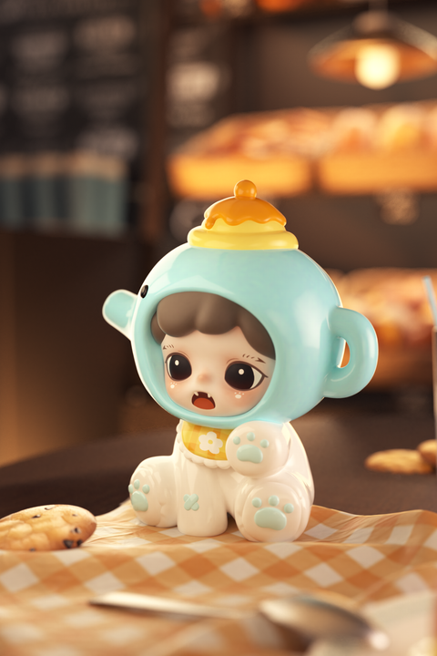 A small figure of a girl in a blue elephant hood sits on an orange checkered napkin. The hood has teapot-shaped ears and a yellow pudding lid on top. She wears white pajamas with paw prints and a yellow bib with a daisy. Cookies and bakery lights fill the background.