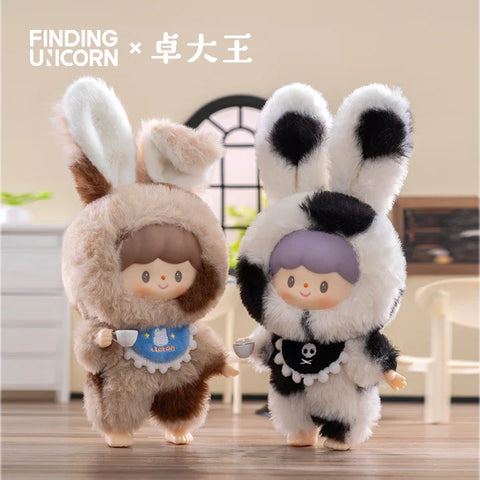 Two plush figures from the Finding Unicorn × 卓大王 Zoton Delicious Bunny Plush Series Blind Box are shown standing in a cozy room setting. The left figure, "Cookie Bunny Zoton," wears a fuzzy brown and cream bunny suit with floppy ears, holding a small teacup and wearing a blue bib that says "Macaron." The right figure, "Oli Bunny Zoton," features a black-and-white spotted bunny suit with a skull emblem on a black bib and holds a matching teacup. Both characters have round, cheerful faces.