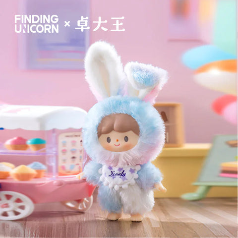 A single plush figure from the Finding Unicorn × 卓大王 Zoton Delicious Bunny Plush Series Blind Box stands in a dreamy pastel setting. This special edition, "Macaron Bunny Zoton," features a soft blend of blue, pink, and white fur with fluffy upright bunny ears. The character wears a ruffled white collar with purple embroidered text that reads "Lovely" and has a sweet, cheerful expression. In the background, a pink dessert cart with colorful cupcakes adds to the whimsical, candy-themed atmosphere.