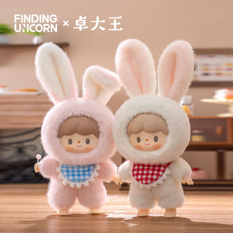 Two plush figures from the Finding Unicorn × 卓大王 Zoton Delicious Bunny Plush Series Blind Box stand side by side in a cozy kitchen-like setting. On the left, "Strawberry Bunny Zoton" wears a fluffy pink bunny suit with large ears and a blue gingham bib, holding a small lollipop. On the right, "Creamy Bunny Zoton" is dressed in a soft white bunny suit with a red gingham bib. Both characters feature matching light brown hair, round faces, and joyful expressions, radiating a sweet and charming vibe.