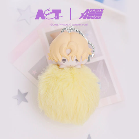 Plush pom-pom keychain depicting Luka from Alien Stage, featuring a blonde chibi character head peeking over a fluffy yellow pom-pom base. The figure includes a silver ball chain and is photographed on a pale background with star-shaped decor elements.