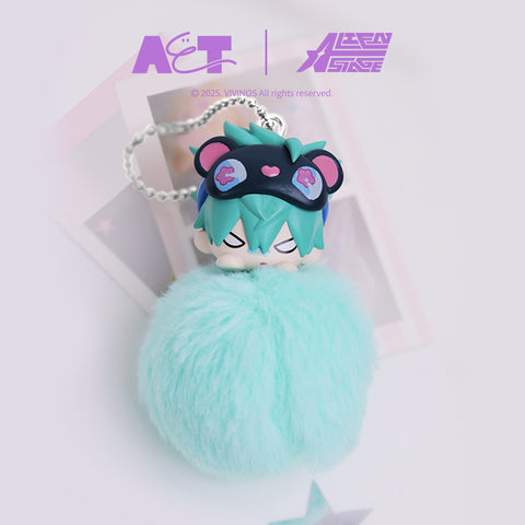Plush keychain figure of Till from Alien Stage with a stylized character head wearing a black sleep mask decorated with pink details. The head sits on a mint green fuzzy pom-pom body with a silver ball chain, shown in a clean studio setting with soft pastel props.
