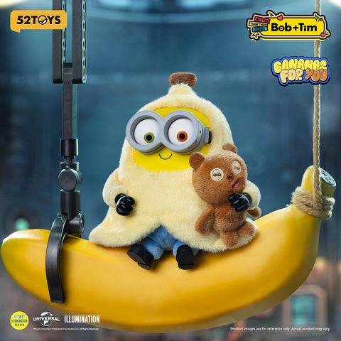 Minion Bob dressed in a yellow banana peel costume, sitting on a large banana swing while holding Tim the bear.