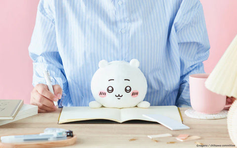 A round white plush with tiny bear-like ears sits on an open notebook in front of a person wearing a blue striped shirt. The plush features embroidered eyes, soft eyebrows, and pink blush marks. A pink mug, a lamp, and stationery items are arranged around the wooden desk in a pastel pink workspace scene.