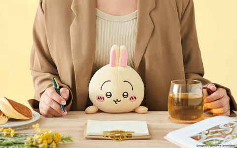 A round beige plush with pink rabbit ears sits on a wooden desk between a person’s hands. The plush has embroidered eyes, eyebrows, a small curved mouth, and pink blush on its cheeks. A cup of tea, a notepad, pancakes, and small yellow flowers surround it in a softly lit, warm-toned scene.