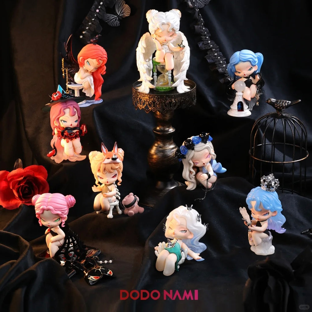 A dark, dramatic display showcases nine gothic fantasy-style chibi figurines arranged on a black velvet backdrop with red roses, lace, butterflies, and a metal birdcage. Each figure has a distinct look, including angel wings, horns, masks, and crystal accessories. The central figure sits elevated with white wings and a golden hourglass. Others feature vibrant hair colors like pink, blue, and red. Below them, red text reads “DODO NAMI”.