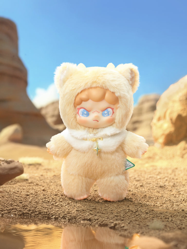 Fluffy cream bear‑hood figure with blue starry eyes and a gold star pendant stands on cracked desert earth. Warm sunlight, eroded stone formations, and a tiny reflective puddle create a sun‑baked canyon backdrop that highlights the toy’s soft textures.
