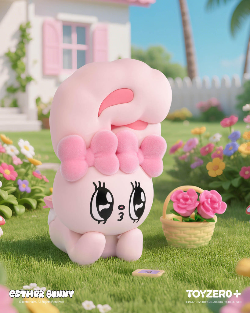 A pastel pink Esther Bunny figurine with large sparkling eyes and a fluffy bow lays adorably on the grass. Next to it is a small basket of pink flowers, surrounded by a garden of colorful blooms, with a white picket fence and cottage-style house in the sunny background.