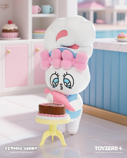 A white Esther Bunny figurine with a pink bow and blue outfit decorates a chocolate cake with pink frosting using a piping bag. The bunny stands beside a small table in a pastel kitchen with cupcakes, mugs, and plates in the background, capturing a sweet and playful baking moment.
