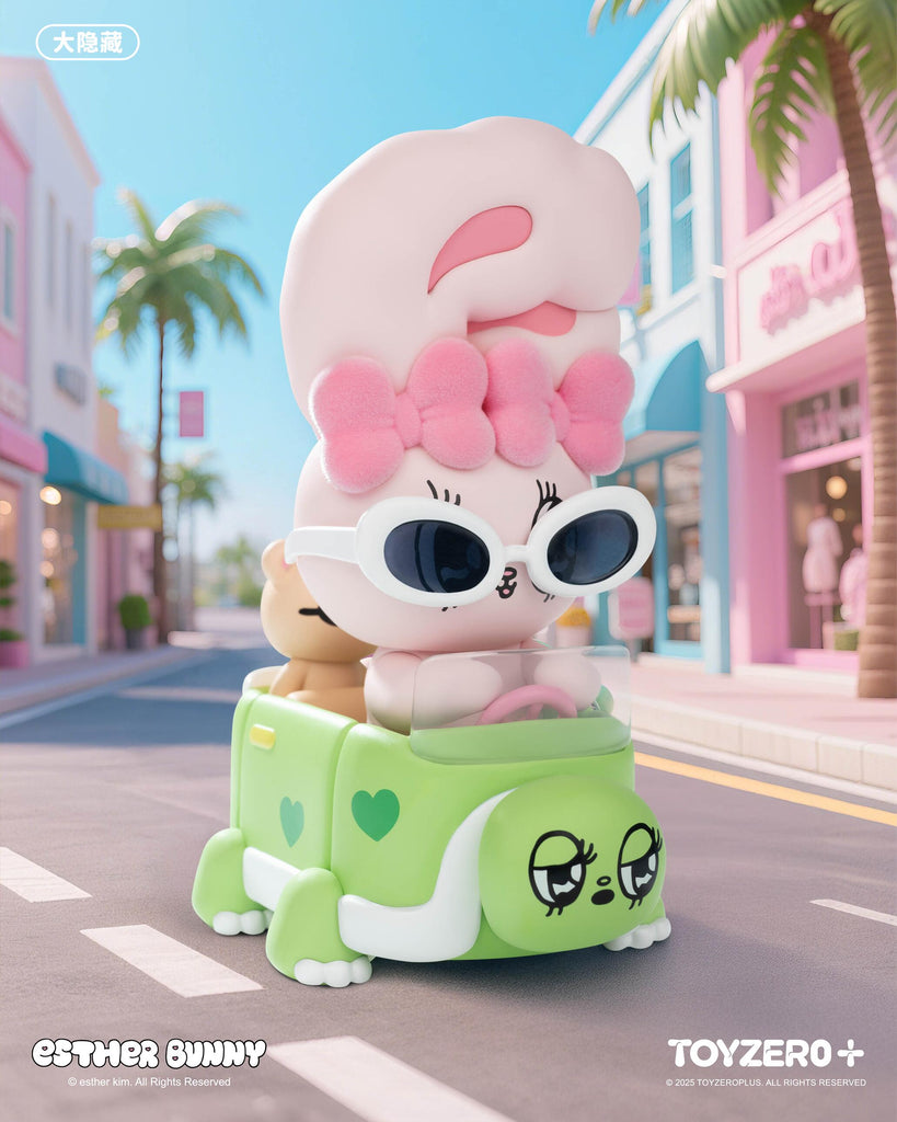 A pastel pink Esther Bunny figurine with a fluffy bow and oversized white sunglasses drives a small green car shaped like a turtle with heart details. A tiny teddy rides along in the backseat as they cruise a colorful street lined with palm trees and pastel pink and blue storefronts.
