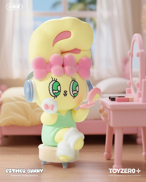 A pastel yellow Esther Bunny figurine with big green eyes and a pink bow sits on a stool in front of a vanity. Wearing a mint outfit, fluffy slippers, and headphones, the bunny holds a compact mirror, playfully applying makeup in a cozy pink bedroom setting.