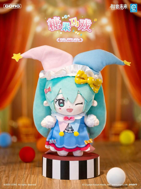 Secret Candy Circus chaser plush shows Hatsune Miku in a pastel clown outfit with blue and pink jester hat, yellow bow, and frilled dress. She winks playfully while posed on a striped stage platform. Background features circus curtains and colorful balls, evoking a whimsical carnival fantasy.