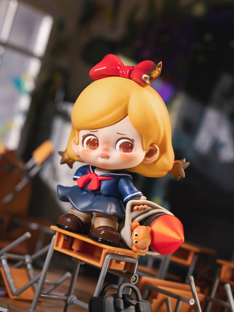 KO Bully figure from the Kokosa KO Girls Series shows a girl in a sailor uniform with red bow, holding a rocket and teddy bear. She stands atop a pile of overturned desks with a worried expression, symbolizing childhood rebellion and playful defiance, contrasted by the chaotic, fiery background details.
